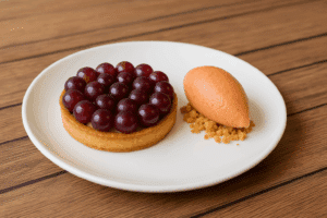 Tarte aux raisins rouges sur une assiette blanche, accompagnée d'un sorbet à l'orange et de crumble sur une table en bois.
