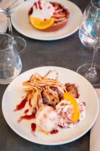 Crêpe revisitée et garnie d'une boule de glace marbrée, de fruits et de sauce rouge, sur une table sombre avec des verres.