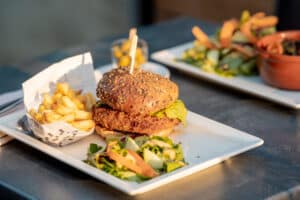 Gros plan sur un burger garni, servi avec des frites dans un papier journal et une petite salade, éclairé par le soleil.