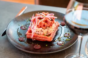 Gaufre salée rouge garnie de salade de chou crémeuse et décorée de fines lamelles de radis rose, sur assiette noire avec vin rouge.