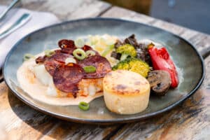 Plat de poisson blanc, chorizo, purée, légumes grillés et flan salé sur une assiette grise posée sur bois.