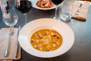 Soupe crémeuse orange avec ravioli et croûtons sur une table sombre, près de verres de vin rouge.