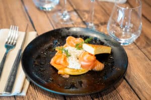 Blinis au saumon fumé et crème fraîche aux herbes, servis sur assiette noire avec couverts et verres sur table en bois.