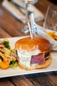 Gros plan d'un burger gourmand avec fromage fondu dégoulinant, jambon sec et salade, servi sur une assiette blanche avec frites en arrière-plan.