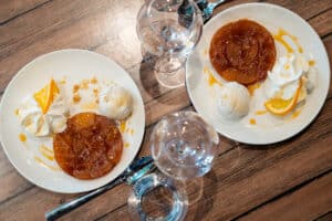 Deux assiettes de Tarte Tatin caramélisée avec une boule de glace vanille, crème fouettée et quartiers d'orange, sur une table en bois, avec des verres à pied.