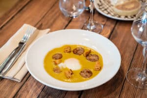 Velouté jaune servi dans une assiette blanche avec un œuf poché, des tranches de chorizo et des croûtons, sur une table en bois.