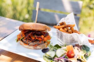 Burger au bacon croustillant, sauce piquante orange, frites maison et salade fraîche servis sur une terrasse.