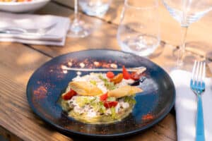 Tartare de crabe et avocat avec herbes fraîches et tomates sur assiette bleue, servi en terrasse ensoleillée.