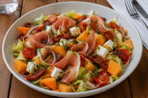 Salade melon, prosciutto et féta avec tomates cerises et pesto dans un bol blanc.