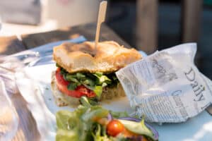 Sandwich focaccia gourmand, tomate, verdure et fromage, accompagné d'une salade fraîche et papier journal.