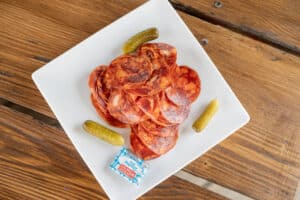 Tranches de chorizo, cornichons et beurre Paysan Breton sur une assiette carrée et table en bois.
