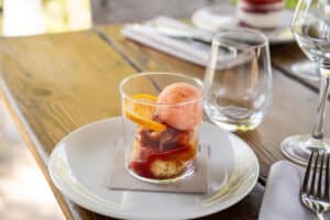 Sorbet rose et dessert aux fruits rouges en verrine avec tranche d'orange sur une table en bois ensoleillée.